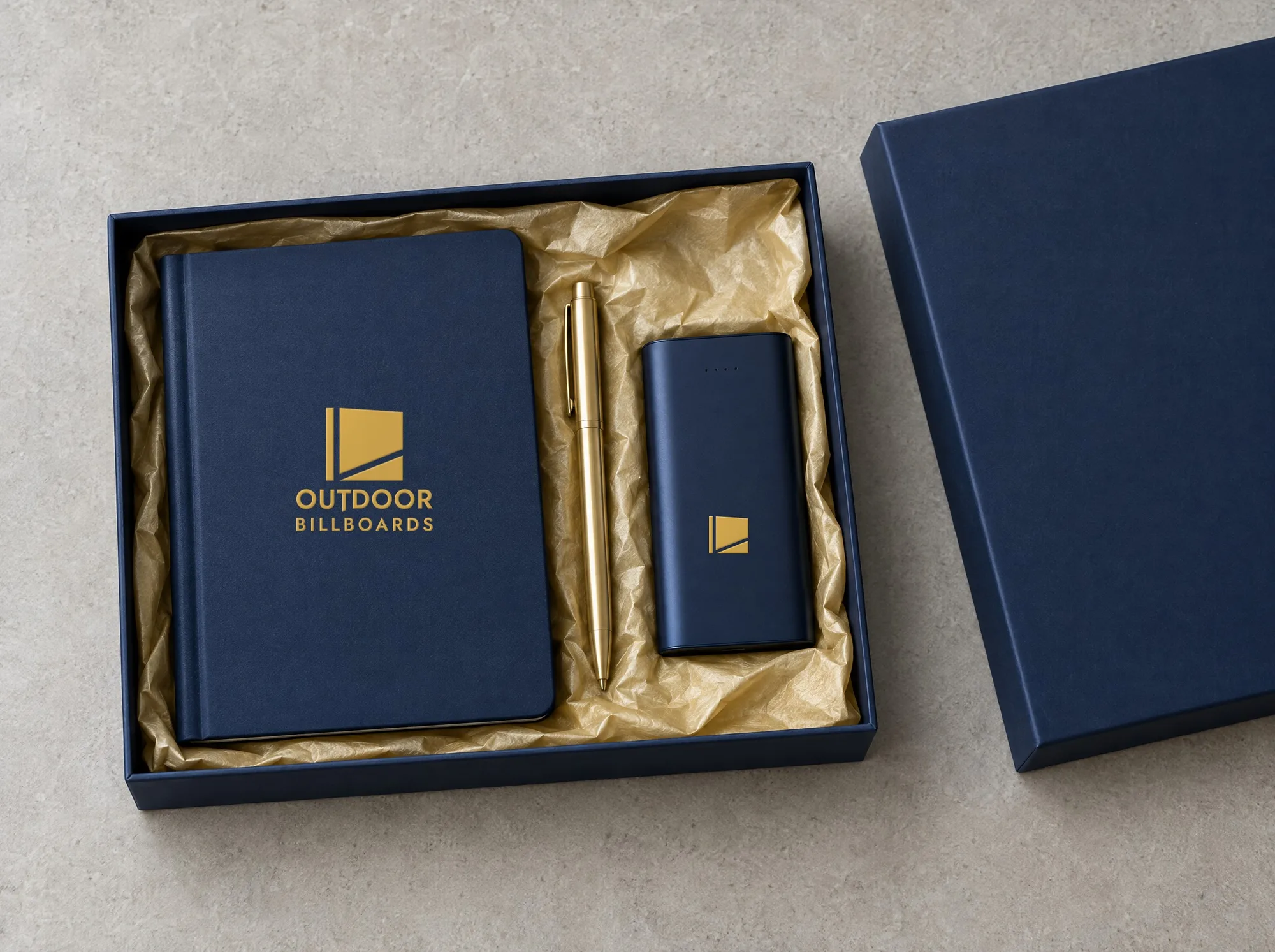 Open navy gift box with a branded notebook, brass pen and power bank on gold tissue