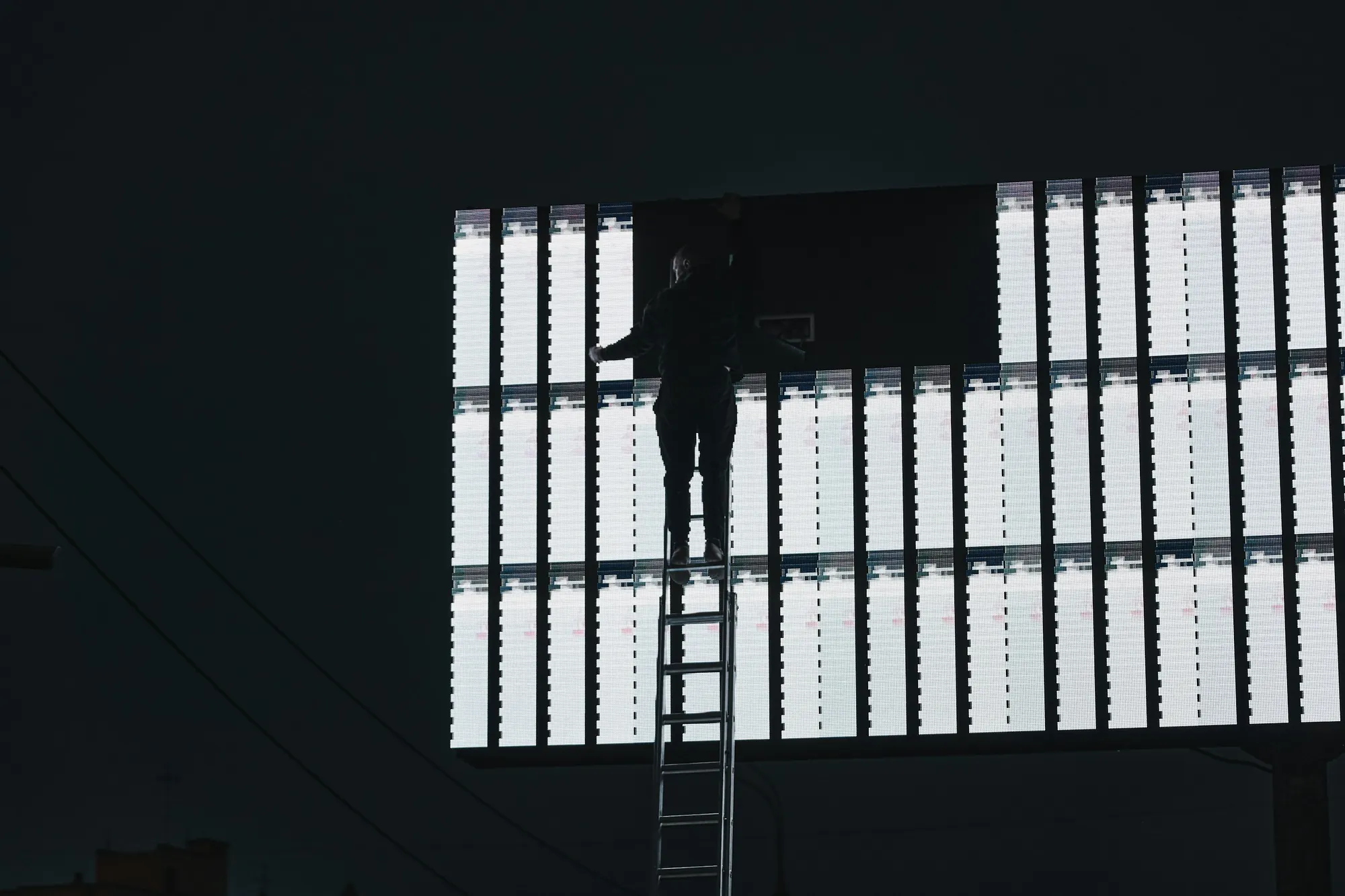 Technician on a ladder fitting panels into a large lit billboard at night